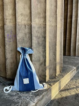 Blue and light denim-colored drawstring fabric bag resting on a stone ledge against a neoclassical column background.
