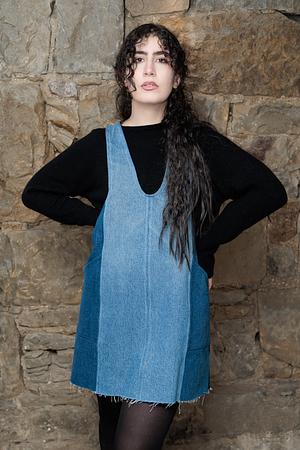 Young woman with long curly hair posing in a denim pinafore dress over a black long-sleeve sweater against a stone wall.
