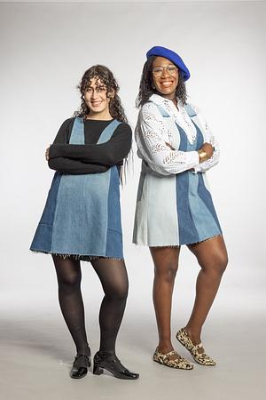 Two smiling women in denim pinefore dresses posing together against a white background.