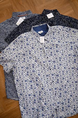 Three short-sleeve men's button-down shirts in blue and white floral patterns laid flat on a wooden surface.